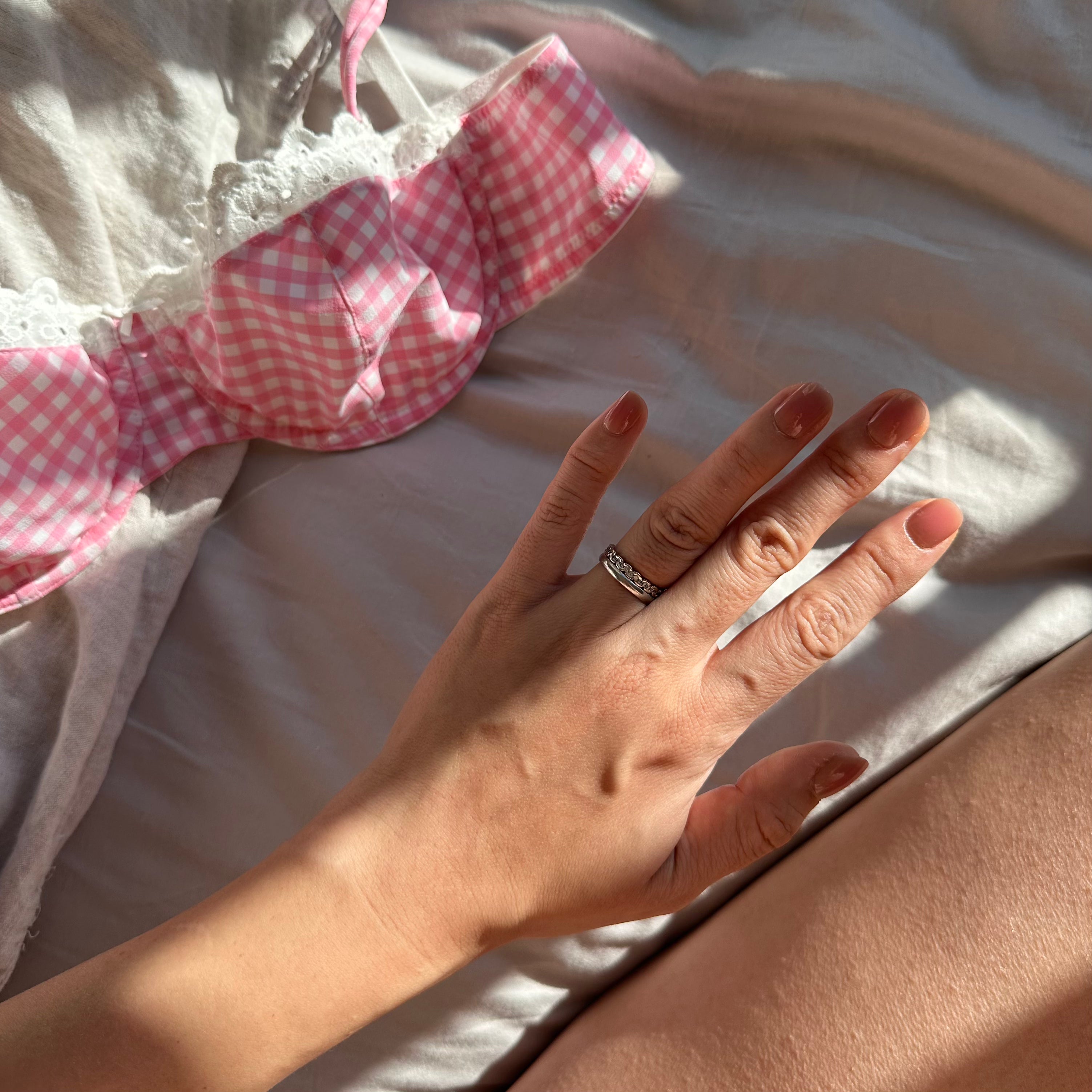Mains avec ongles manucurés posées sur un tissu brodé, ruban à carreaux roses en arrière-plan